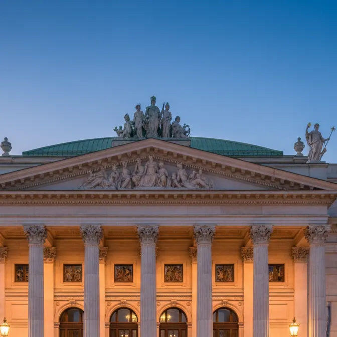 Columns, sculpted pediment and rooftop statues of a floodlit neoclassical facade at dusk
